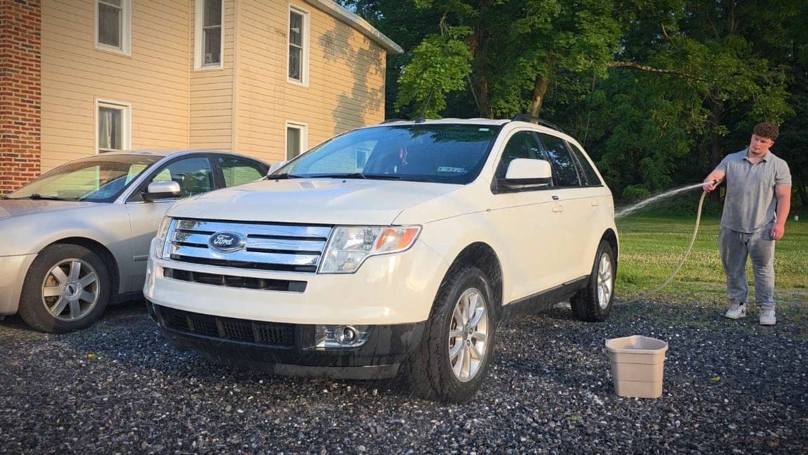 White Ford Edge being rinsed with a hose in a gravel driveway during an exterior wash detail