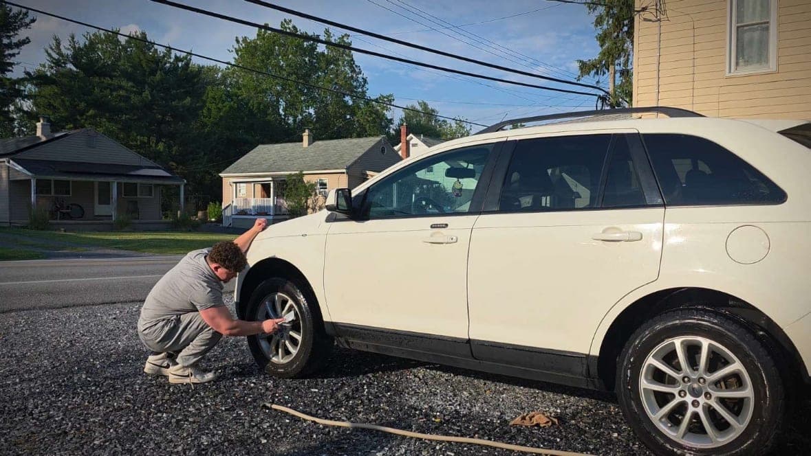 Mike crouched down, hand-scrubbing a front wheel and tire on a white Ford Edge in a gravel driveway